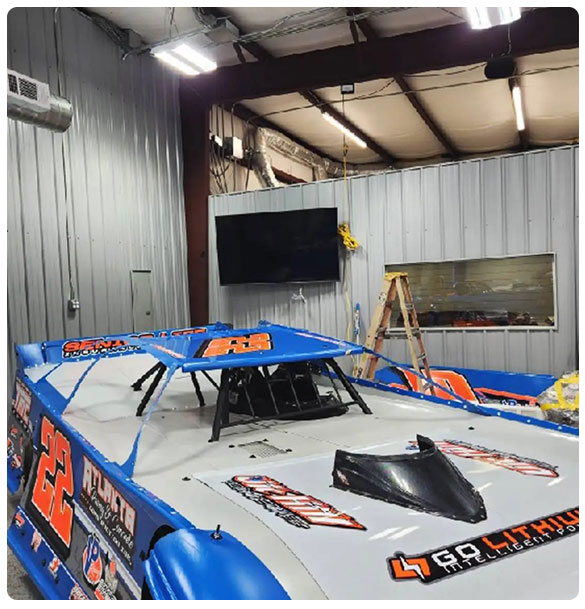 Motorsport workshop interior featuring a blue race car, mounted TV for team briefings, and ladder for installation, illustrating SAV Systems' project for advanced audio and video solutions.