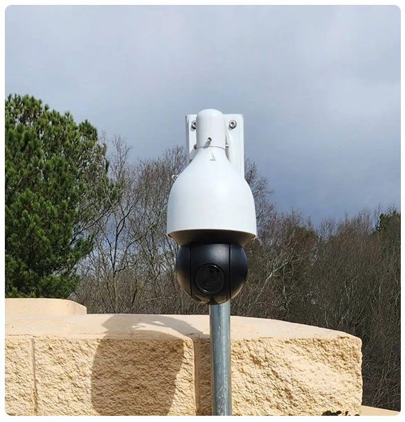 Surveillance camera mounted on a pole, part of Standridge Color Corp's extensive security system installation, with a natural backdrop and cloudy sky.