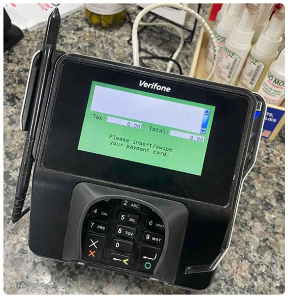 Payment terminal displaying prompts for card insertion/swiping, indicating tax and total amounts as zero, positioned on a countertop with various products in the background.